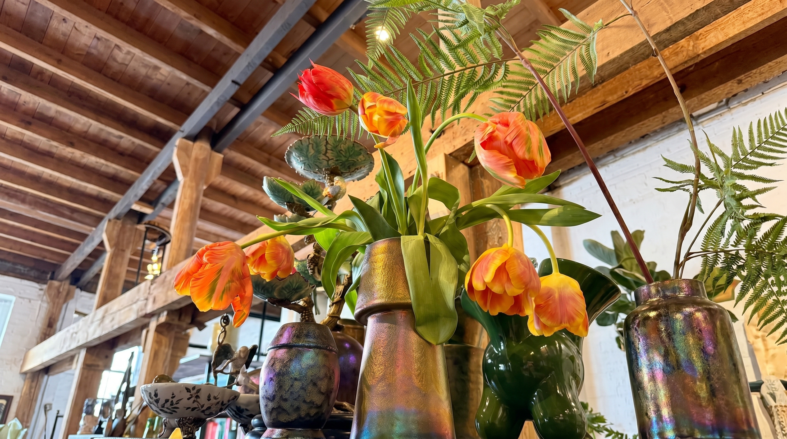 Bloemenarrangement met oranje tulpen en varens in geïriseerde vazen onder houten plafondbalken bij Nozili.